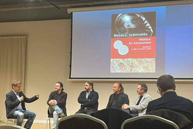 A könyvbemutató előadói egy színpadon ülnek, a háttérben a könyv borítójának képe látható kivetítve.